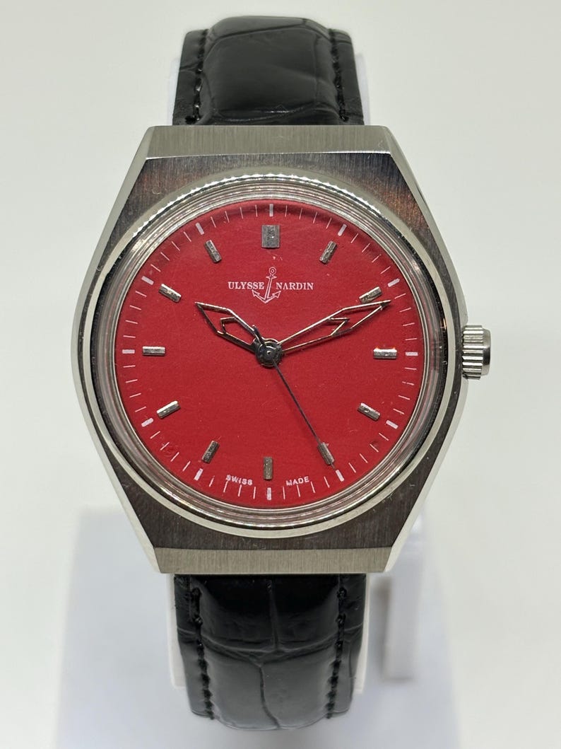 Puede incluir: Un reloj de pulsera Ulysse Nardin con esfera roja vibrante, marcadores de hora plateados y manecillas caladas. El reloj cuenta con una caja de acero inoxidable cepillado y una correa de cuero negro con un patr&oacute;n texturizado de cocodrilo.