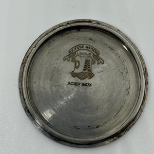 K&ouml;nnte beinhalten: Ein kreisf&ouml;rmiger Uhrengeh&auml;useboden aus silberfarbenem Metall mit eingraviertem Ulysse Nardin-Logo sowie dem Schriftzug Le Locle und Acier Inox. Die Metalloberfl&auml;che weist sichtbare Gebrauchsspuren und Oxidation am Rand auf.