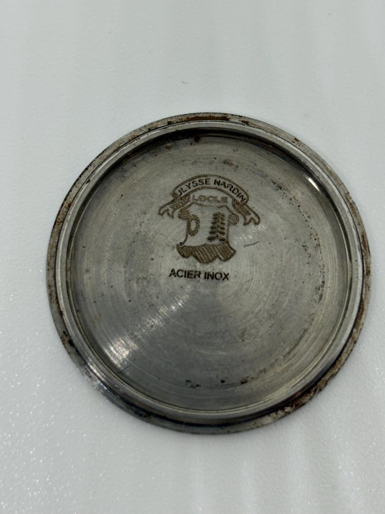 K&ouml;nnte beinhalten: Ein kreisf&ouml;rmiger Uhrengeh&auml;useboden aus silberfarbenem Metall mit eingraviertem Ulysse Nardin-Logo sowie dem Schriftzug Le Locle und Acier Inox. Die Metalloberfl&auml;che weist sichtbare Gebrauchsspuren und Oxidation am Rand auf.