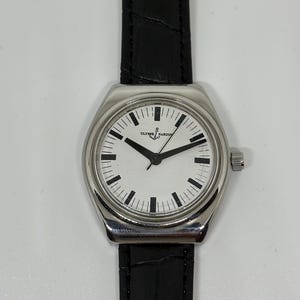 May include: A wristwatch with a silver-toned stainless steel case and a white dial featuring black hour markers and hands. The dial displays the text ULYSSE NARDIN with an anchor logo. It is attached to a black leather strap with a textured finish.
