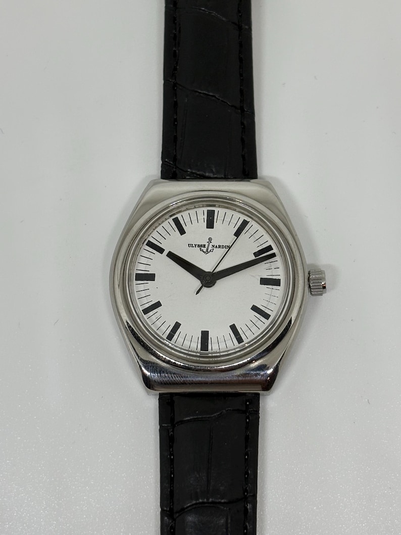 May include: A wristwatch with a silver-toned stainless steel case and a white dial featuring black hour markers and hands. The dial displays the text ULYSSE NARDIN with an anchor logo. It is attached to a black leather strap with a textured finish.