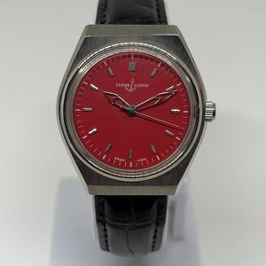 Puede incluir: Un reloj de pulsera Ulysse Nardin con una esfera roja vibrante, marcadores de hora y manecillas en tono plateado. El reloj tiene una caja de acero inoxidable cepillado y una correa de cuero negro con patr&oacute;n de cocodrilo. Se lee Ulysse Nardin.