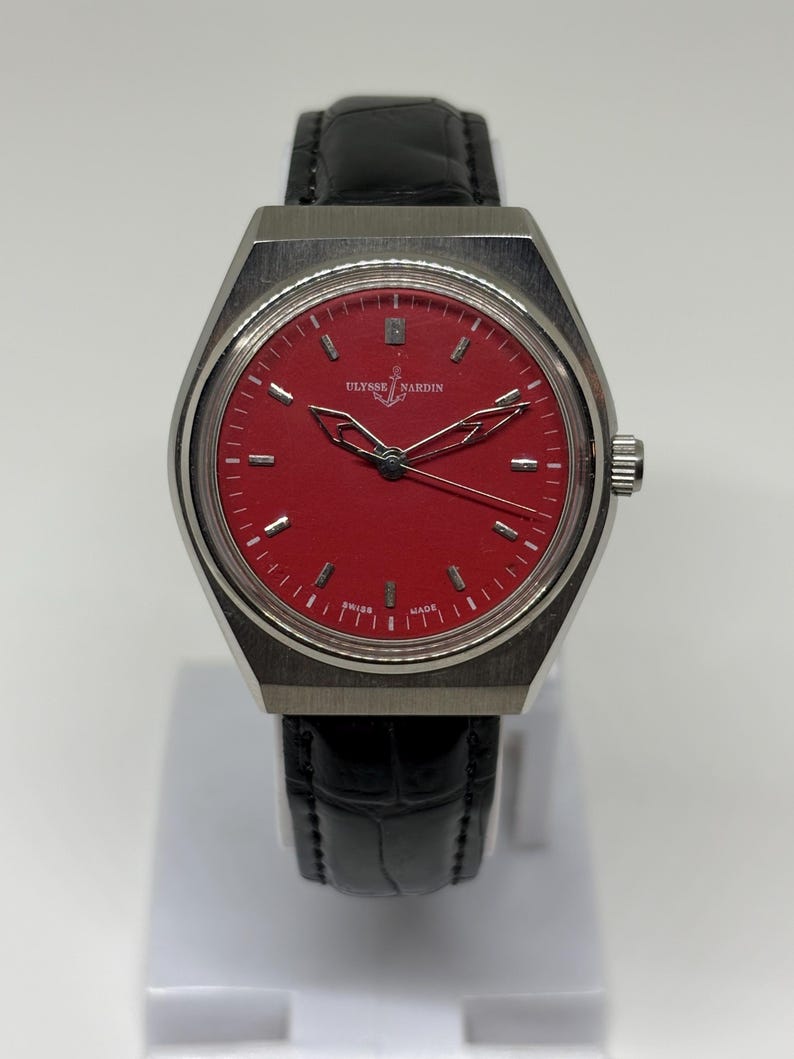 Puede incluir: Un reloj de pulsera Ulysse Nardin con una esfera roja vibrante, marcadores de hora y manecillas en tono plateado. El reloj tiene una caja de acero inoxidable cepillado y una correa de cuero negro con patr&oacute;n de cocodrilo. Se lee Ulysse Nardin.