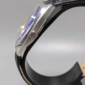 May include: A silver-toned wristwatch with a black leather band. The watch face has a blue and silver design. The watch is displayed on a black stand. The watch has a small emblem on the side.