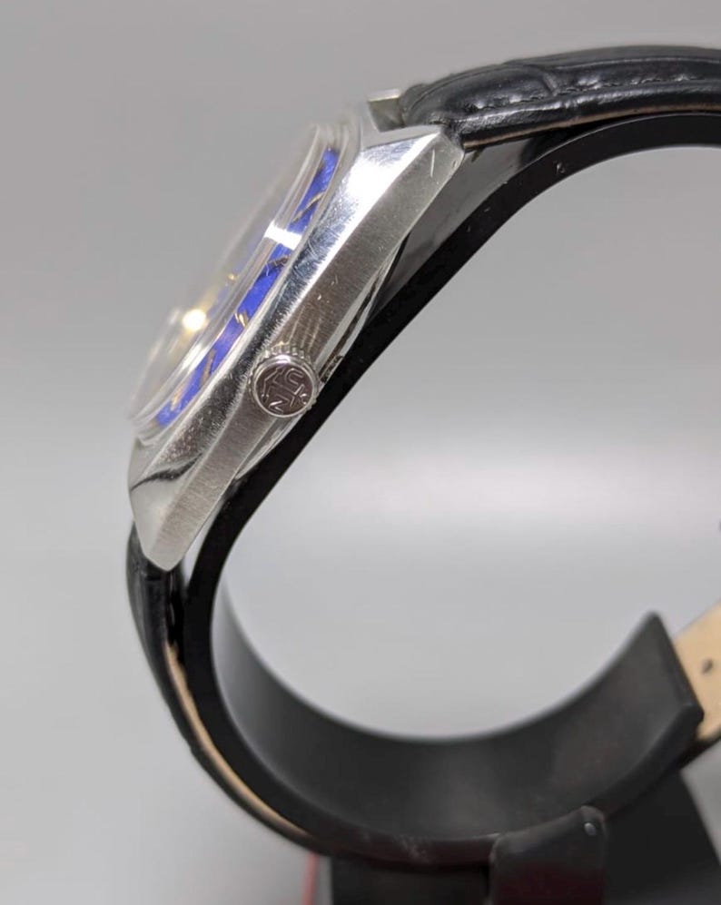 May include: A silver-toned wristwatch with a black leather band. The watch face has a blue and silver design. The watch is displayed on a black stand. The watch has a small emblem on the side.