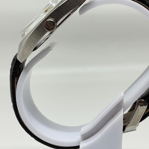 K&ouml;nnte beinhalten: Eine Seitenansicht einer silberfarbenen Armbanduhr mit einem strukturierten schwarzen Lederarmband mit Krokodilmuster. Das Uhrengeh&auml;use ist metallisch und hat eine runde Krone an der Seite, die ein kleines Logo zeigt. Die Uhr steht auf einem wei&szlig;en St&auml;nder.