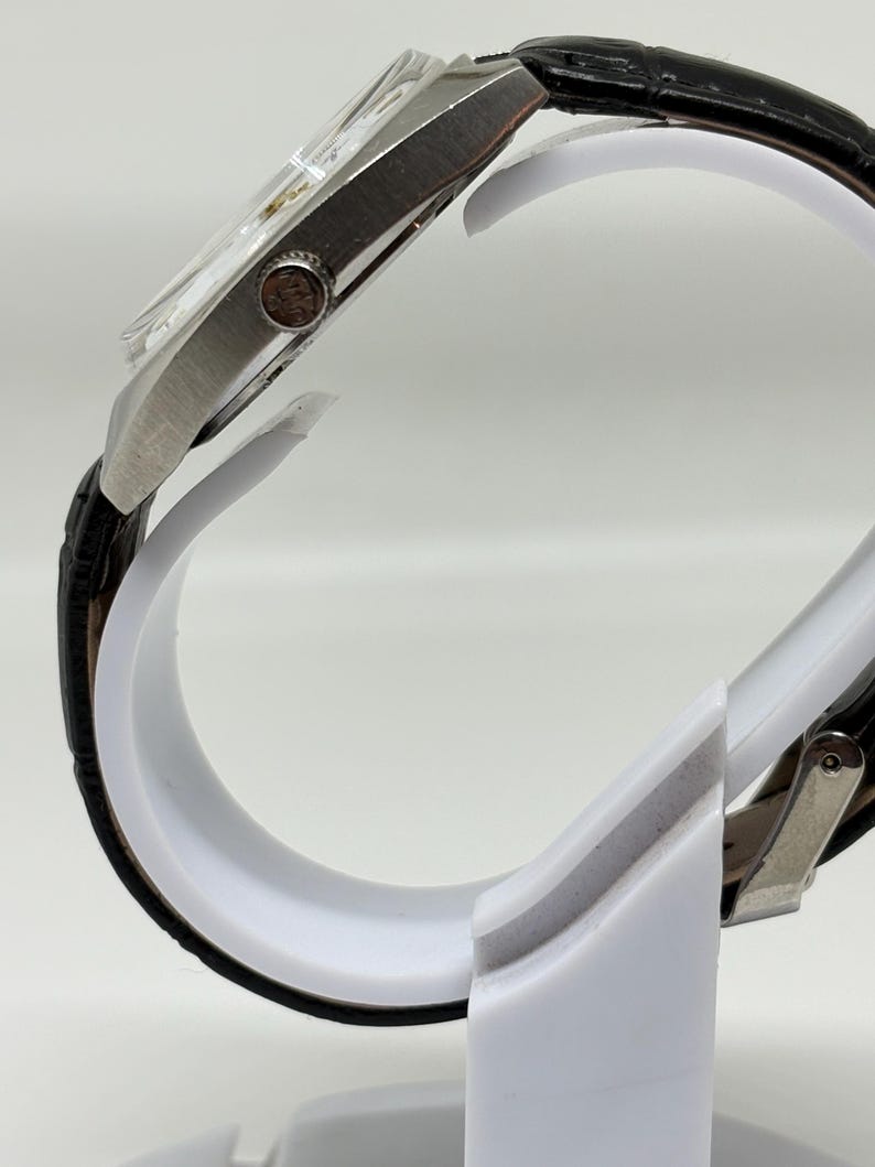 K&ouml;nnte beinhalten: Eine Seitenansicht einer silberfarbenen Armbanduhr mit einem strukturierten schwarzen Lederarmband mit Krokodilmuster. Das Uhrengeh&auml;use ist metallisch und hat eine runde Krone an der Seite, die ein kleines Logo zeigt. Die Uhr steht auf einem wei&szlig;en St&auml;nder.