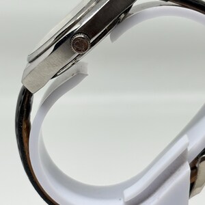 May include: A side view of a wristwatch featuring a silver-toned metal case with a textured crown and a black leather strap with a visible grain pattern. The watch case has a polished finish and is mounted on a white display stand.