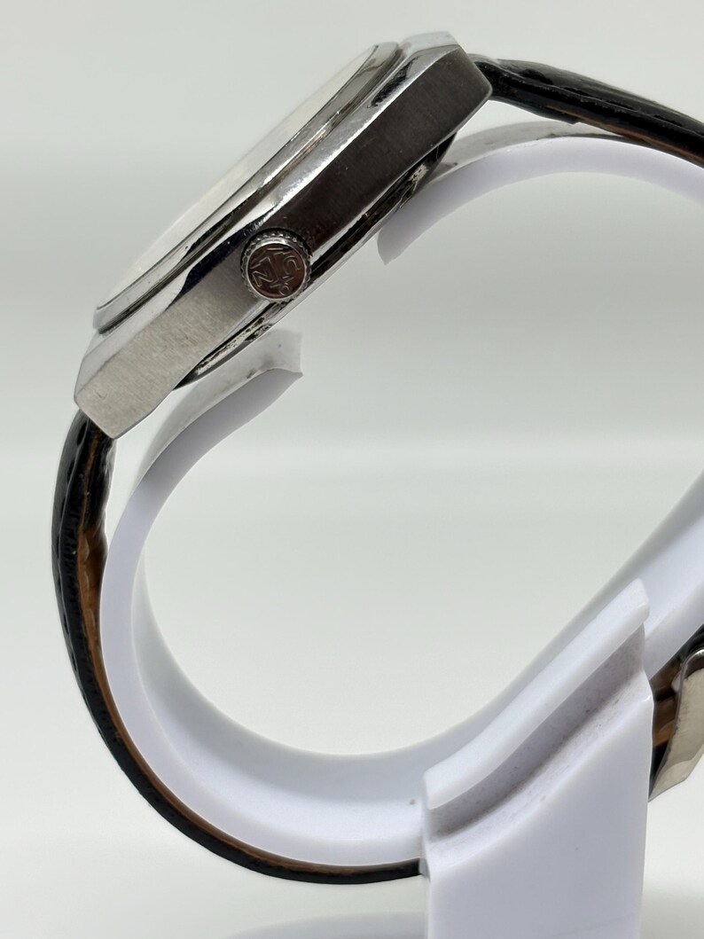 May include: A side view of a wristwatch featuring a silver-toned metal case with a textured crown and a black leather strap with a visible grain pattern. The watch case has a polished finish and is mounted on a white display stand.