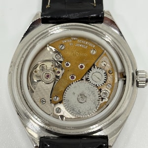 May include: A silver-toned wristwatch with the back casing removed to reveal the internal mechanical movement. The movement features a gold-colored bridge engraved with "SWISS SEVENTEEN 17 JEWELS" and "ULYSSE NARDIN," alongside visible gears.