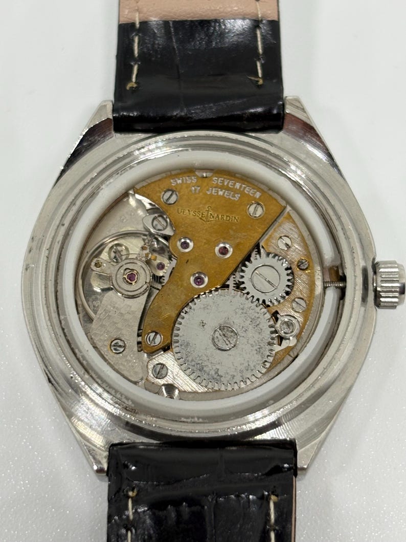 May include: A silver-toned wristwatch with the back casing removed to reveal the internal mechanical movement. The movement features a gold-colored bridge engraved with "SWISS SEVENTEEN 17 JEWELS" and "ULYSSE NARDIN," alongside visible gears.