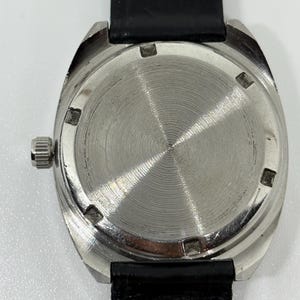 May include: A silver-toned watch case with a circular, brushed metal back featuring six notches around the perimeter. A small, ridged silver crown is positioned on the left side of the case. The watch is attached to a black textured leather strap.