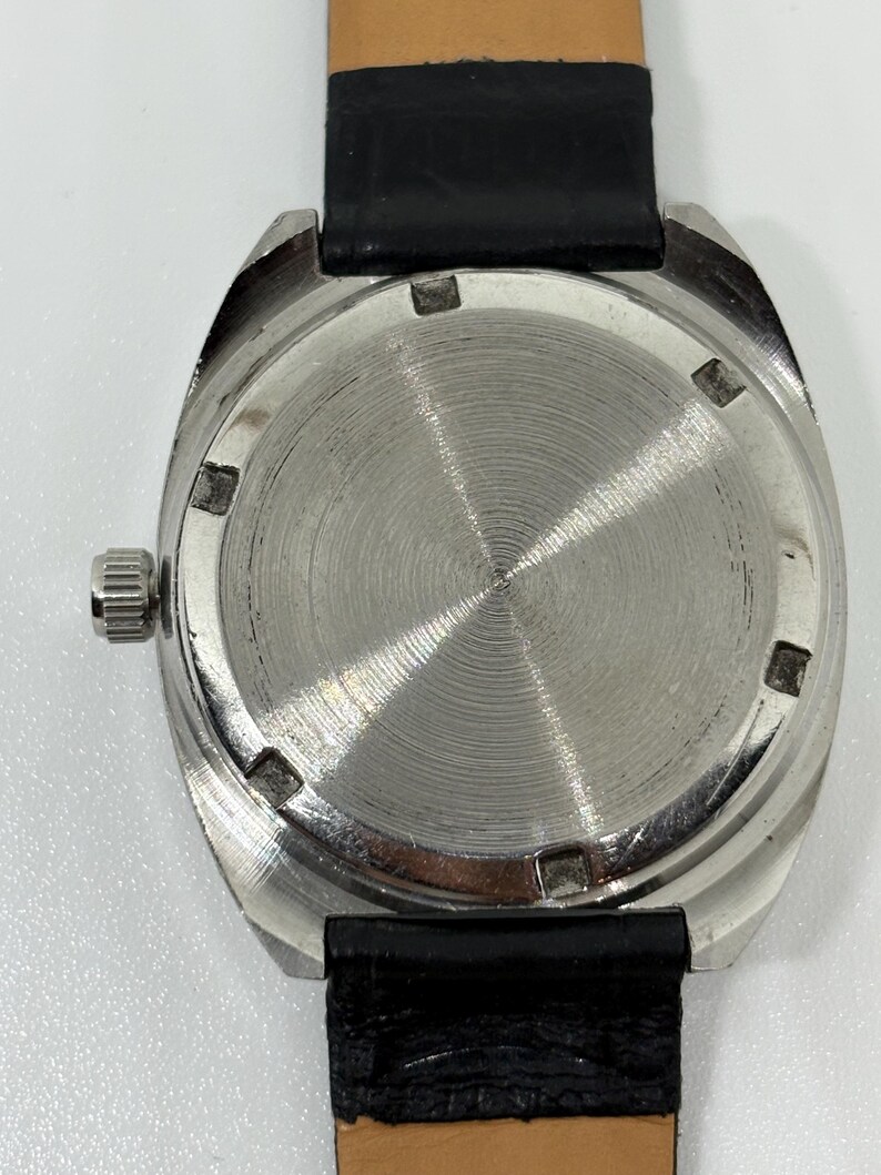 May include: A silver-toned watch case with a circular, brushed metal back featuring six notches around the perimeter. A small, ridged silver crown is positioned on the left side of the case. The watch is attached to a black textured leather strap.