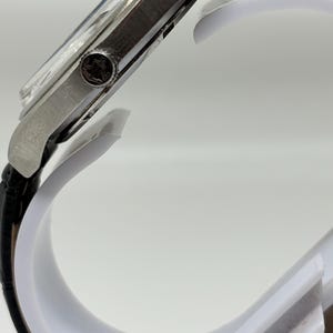 May include: A silver-toned wristwatch with a black leather strap. The side profile shows a metallic case with a circular crown featuring an engraved logo. The watch face is partially visible under a clear crystal, and the strap has a textured finish.