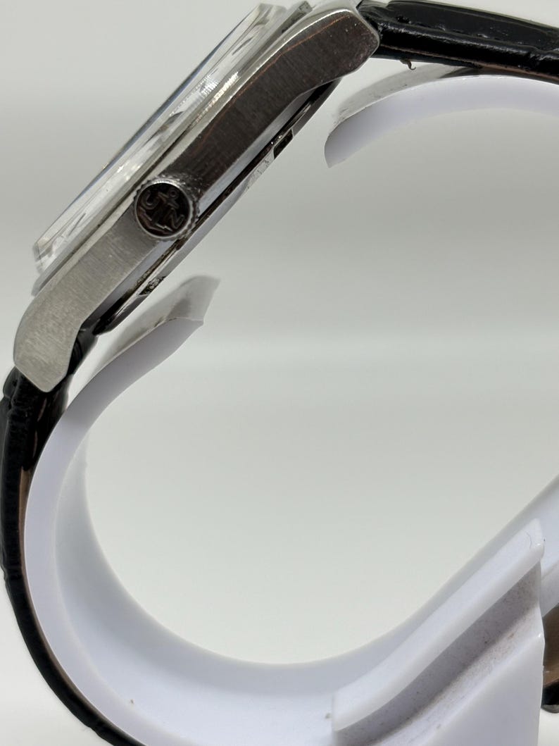 May include: A silver-toned wristwatch with a black leather strap. The side profile shows a metallic case with a circular crown featuring an engraved logo. The watch face is partially visible under a clear crystal, and the strap has a textured finish.