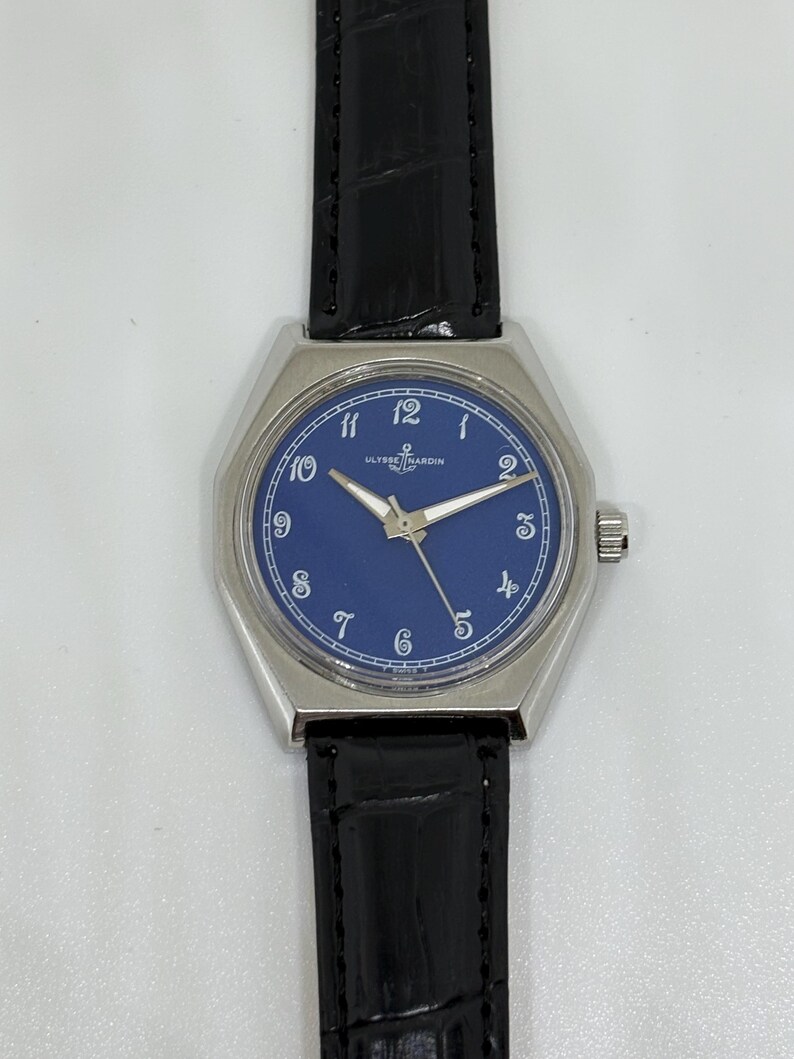 May include: A wristwatch with a silver-toned octagonal case and a deep blue dial featuring white Arabic numerals. The watch has silver-toned hands and a black leather strap with a textured finish. The Ulysse Nardin logo is printed on the dial.