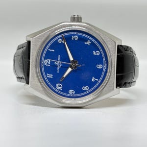 May include: A wristwatch with a brushed silver-tone octagonal case and a vibrant blue dial. The dial features white Arabic numerals, silver-tone hands, and the Ulysse Nardin logo with an anchor symbol. It has a black leather strap with a textured finish.