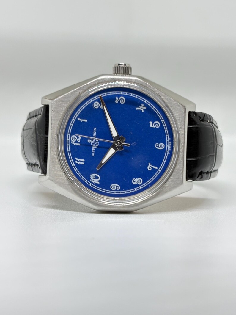 May include: A wristwatch with a brushed silver-tone octagonal case and a vibrant blue dial. The dial features white Arabic numerals, silver-tone hands, and the Ulysse Nardin logo with an anchor symbol. It has a black leather strap with a textured finish.