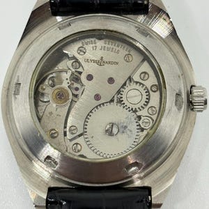 Puede incluir: Un reloj de pulsera plateado con una tapa trasera transparente que revela el movimiento mec&aacute;nico. El mecanismo est&aacute; grabado con 'SWISS', 'SEVENTEEN 17 JEWELS' y 'ULYSSE NARDIN'. Cuenta con una correa de cuero negro con costuras blancas visibles.