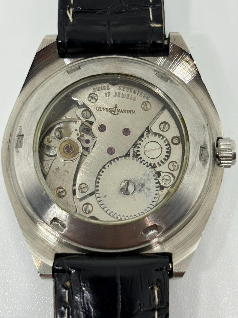 Puede incluir: Un reloj de pulsera plateado con una tapa trasera transparente que revela el movimiento mec&aacute;nico. El mecanismo est&aacute; grabado con 'SWISS', 'SEVENTEEN 17 JEWELS' y 'ULYSSE NARDIN'. Cuenta con una correa de cuero negro con costuras blancas visibles.
