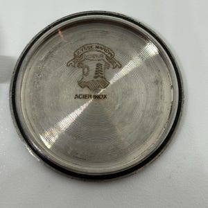 May include: A circular silver-toned metal watch case back featuring a concentric circular brushed texture. The center displays an engraved logo with the text Ulysse Nardin and a shield emblem, with the words ACIER INOX printed below the emblem.