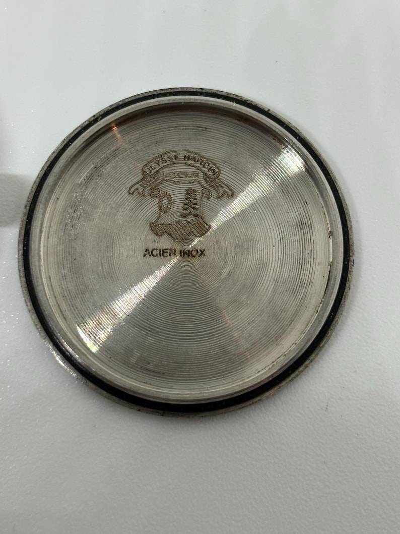 May include: A circular silver-toned metal watch case back featuring a concentric circular brushed texture. The center displays an engraved logo with the text Ulysse Nardin and a shield emblem, with the words ACIER INOX printed below the emblem.