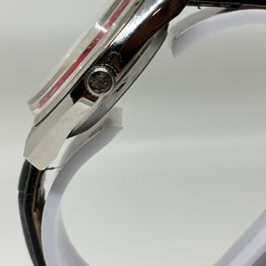 Puede incluir: Una vista lateral de un reloj de pulsera con caja de metal plateado pulido y correa de cuero negro. El reloj presenta una corona de cuerda prominente en el lateral con un logotipo circular grabado y un acento rojo visible bajo el cristal.