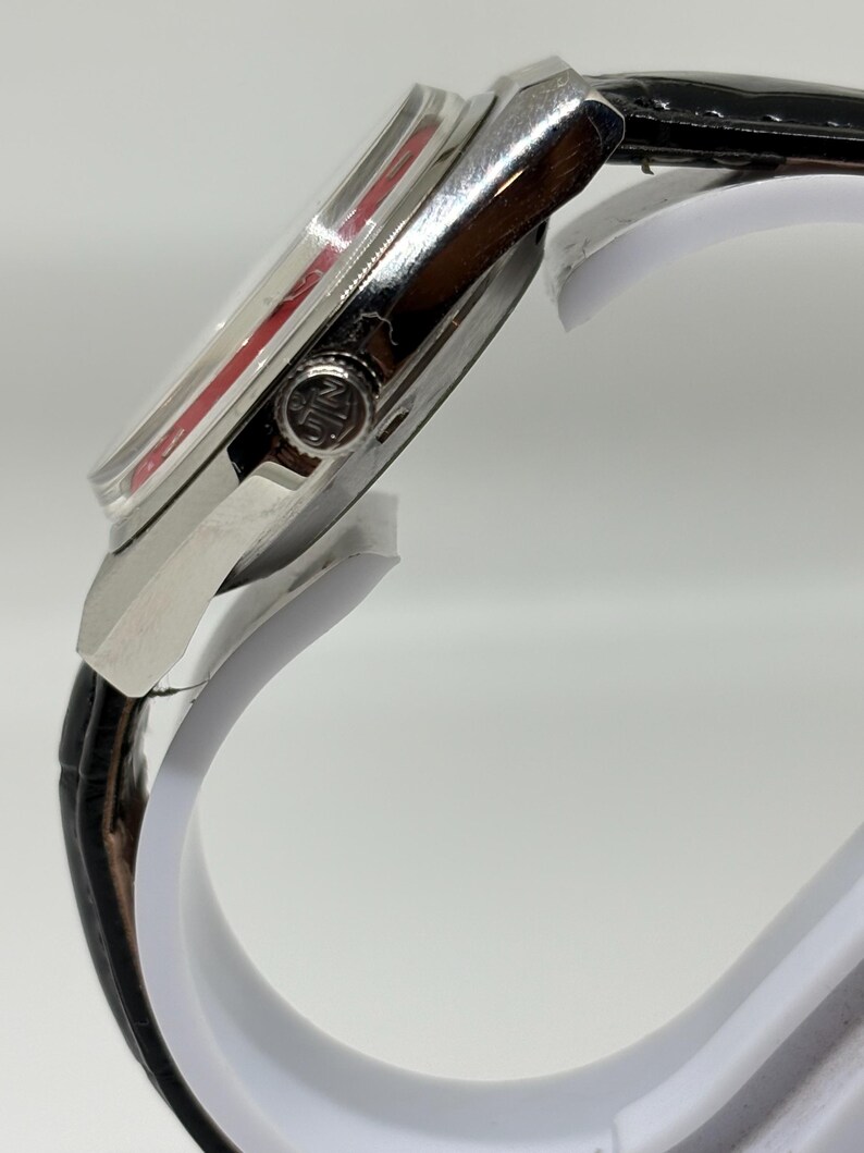 Puede incluir: Una vista lateral de un reloj de pulsera con caja de metal plateado pulido y correa de cuero negro. El reloj presenta una corona de cuerda prominente en el lateral con un logotipo circular grabado y un acento rojo visible bajo el cristal.