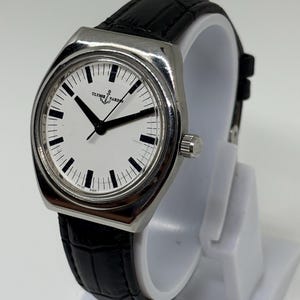 May include: A silver-tone Ulysse Nardin wristwatch with a white dial, black hour markers, and black hands. The watch features a black leather strap with a textured pattern and a silver crown on the side. The dial displays the Ulysse Nardin logo.