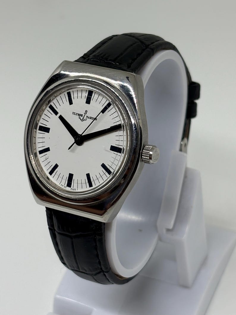 May include: A silver-tone Ulysse Nardin wristwatch with a white dial, black hour markers, and black hands. The watch features a black leather strap with a textured pattern and a silver crown on the side. The dial displays the Ulysse Nardin logo.