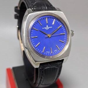May include: A vintage Ulysse Nardin wristwatch with a striking blue dial and gold-tone hands and markers. The watch features a stainless steel case and a black leather strap. The watch is displayed on a black and red stand.