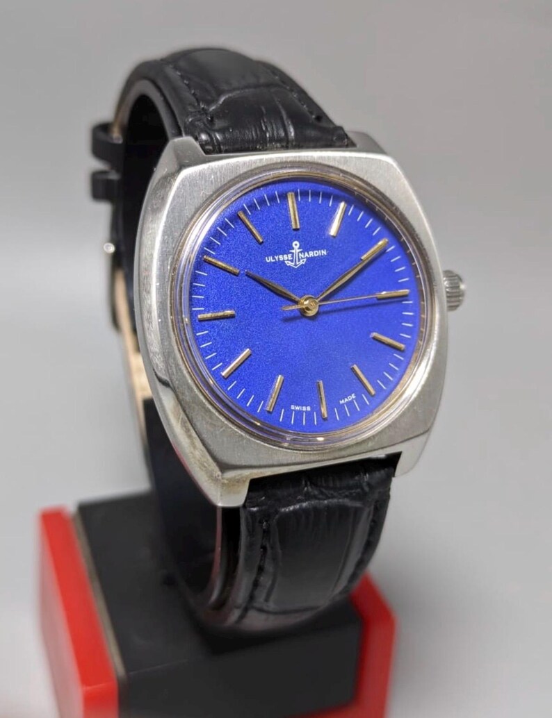 May include: A vintage Ulysse Nardin wristwatch with a striking blue dial and gold-tone hands and markers. The watch features a stainless steel case and a black leather strap. The watch is displayed on a black and red stand.