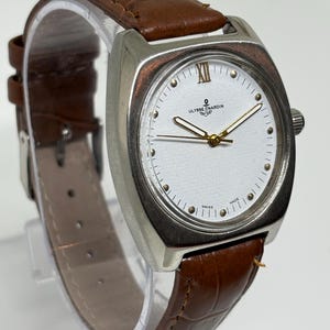 K&ouml;nnte beinhalten: Eine Ulysse Nardin Armbanduhr mit einem silberfarbenen kissenf&ouml;rmigen Geh&auml;use, einem wei&szlig;en Zifferblatt mit goldfarbenen Zeigern, einer r&ouml;mischen XII oben und einem braunen Lederarmband mit Krokodilpr&auml;gung und einer Metallschnalle.