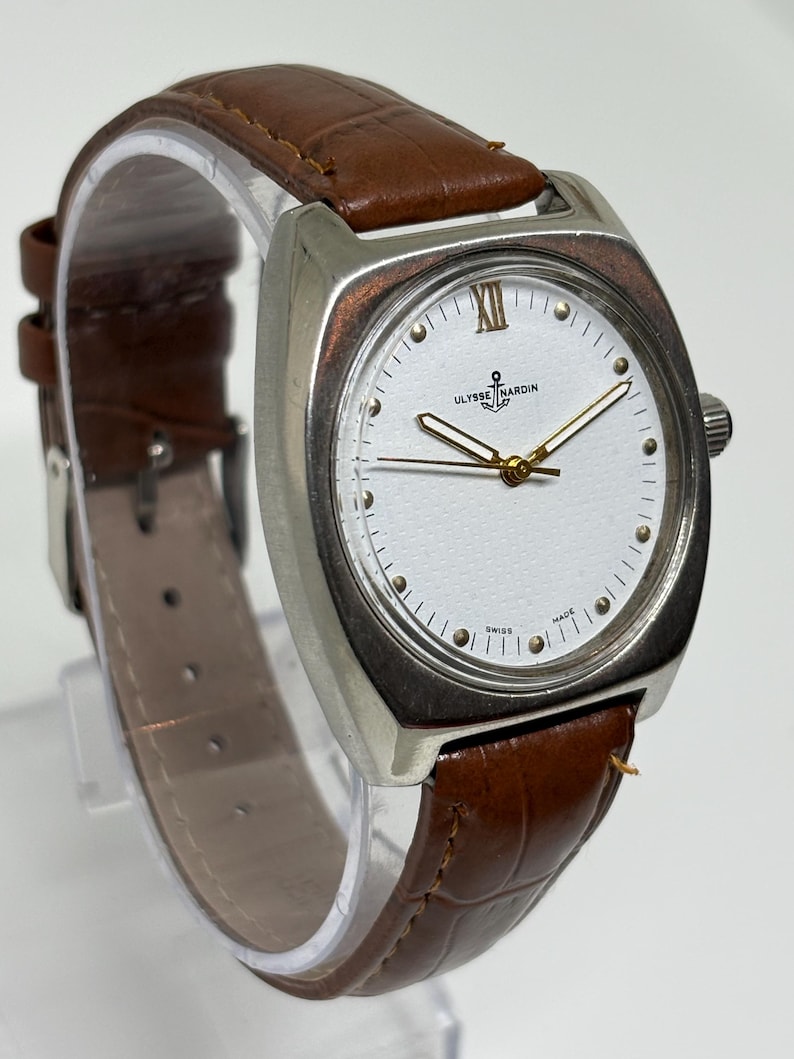 K&ouml;nnte beinhalten: Eine Ulysse Nardin Armbanduhr mit einem silberfarbenen kissenf&ouml;rmigen Geh&auml;use, einem wei&szlig;en Zifferblatt mit goldfarbenen Zeigern, einer r&ouml;mischen XII oben und einem braunen Lederarmband mit Krokodilpr&auml;gung und einer Metallschnalle.