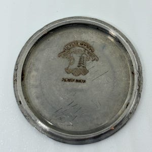 May include: A circular silver-toned metal watch case back featuring an engraved Ulysse Nardin logo at the top and the text ACIER INOX printed below. The metal surface shows signs of wear with faint scratches and a raised outer rim edge.