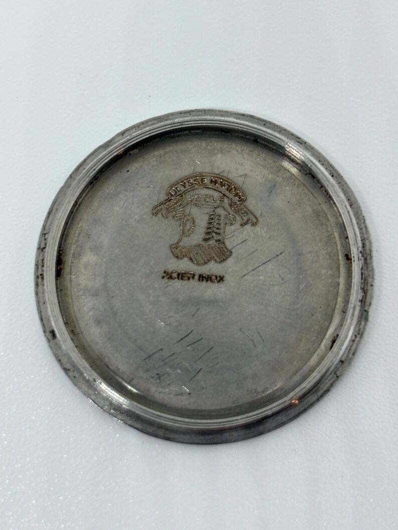 May include: A circular silver-toned metal watch case back featuring an engraved Ulysse Nardin logo at the top and the text ACIER INOX printed below. The metal surface shows signs of wear with faint scratches and a raised outer rim edge.
