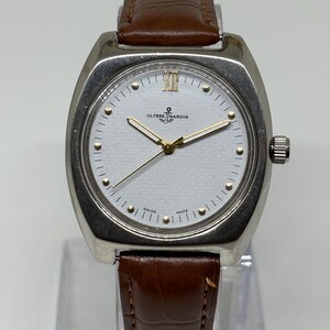 K&ouml;nnte beinhalten: Eine silberfarbene Armbanduhr mit wei&szlig;em, strukturiertem Zifferblatt, goldfarbenen Zeigern und goldfarbenen Stundenmarkierungen. Die Uhr zeigt das Ulysse Nardin-Logo und ein Ankersymbol bei zw&ouml;lf Uhr sowie den Schriftzug SWISS MADE unten.