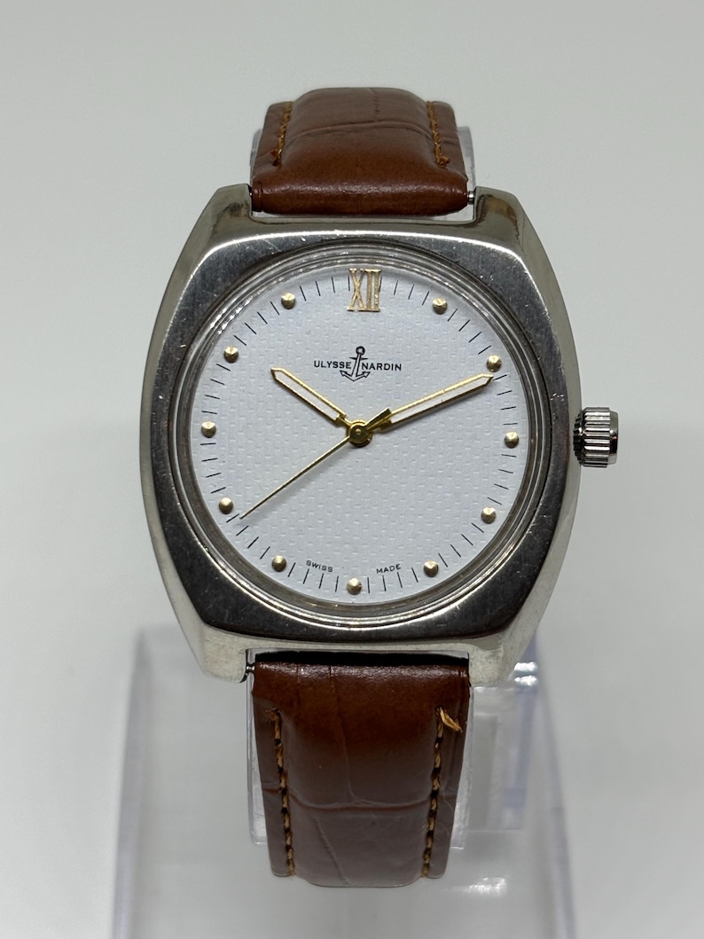 K&ouml;nnte beinhalten: Eine silberfarbene Armbanduhr mit wei&szlig;em, strukturiertem Zifferblatt, goldfarbenen Zeigern und goldfarbenen Stundenmarkierungen. Die Uhr zeigt das Ulysse Nardin-Logo und ein Ankersymbol bei zw&ouml;lf Uhr sowie den Schriftzug SWISS MADE unten.