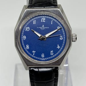 May include: A wristwatch with a blue dial, white Arabic numerals, and silver-tone hands. The watch features a silver-tone metal case and a black leather strap with a textured finish. The text ULYSSE NARDIN and an anchor logo are printed on the dial.
