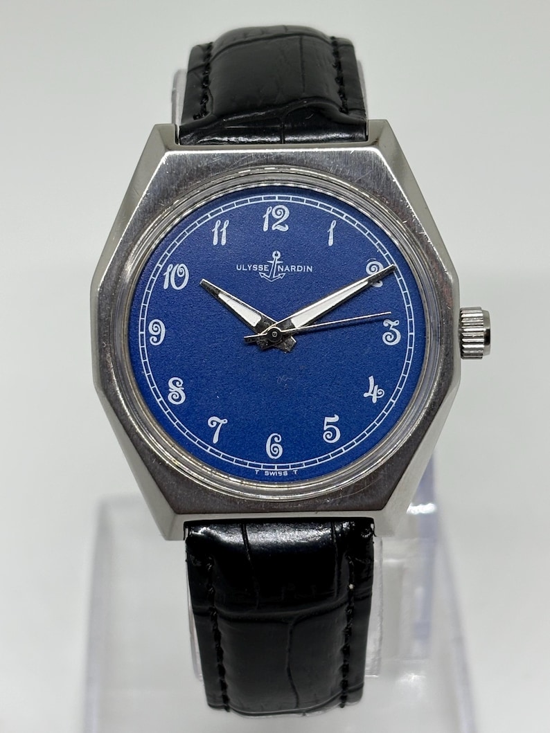 May include: A wristwatch with a blue dial, white Arabic numerals, and silver-tone hands. The watch features a silver-tone metal case and a black leather strap with a textured finish. The text ULYSSE NARDIN and an anchor logo are printed on the dial.