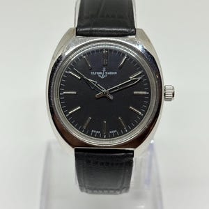 May include: A Ulysse Nardin wristwatch featuring a round silver-tone case, a black dial with silver-tone hour markers, and silver-tone hands. The watch includes a black leather strap with a textured finish and a silver-tone crown on the side.