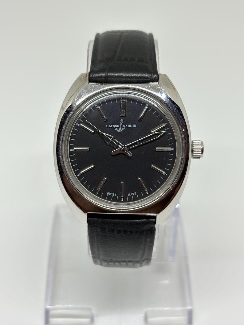 May include: A Ulysse Nardin wristwatch featuring a round silver-tone case, a black dial with silver-tone hour markers, and silver-tone hands. The watch includes a black leather strap with a textured finish and a silver-tone crown on the side.