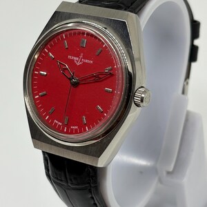 Puede incluir: Un reloj de pulsera con esfera circular roja, marcadores de hora plateados y manecillas plateadas. El reloj cuenta con una caja de metal plateado cepillado y una correa de cuero negro texturizado. La esfera muestra el texto Ulysse Nardin y Swiss Made.