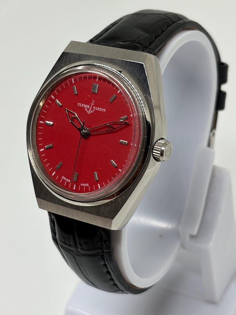 Puede incluir: Un reloj de pulsera con esfera circular roja, marcadores de hora plateados y manecillas plateadas. El reloj cuenta con una caja de metal plateado cepillado y una correa de cuero negro texturizado. La esfera muestra el texto Ulysse Nardin y Swiss Made.