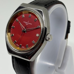 May include: A Ulysse Nardin wristwatch featuring a vibrant red dial with silver-tone hour markers and hands. The watch has a polished silver-tone metal case, a textured black leather strap, and the text Ulysse Nardin and Swiss Made on the dial.
