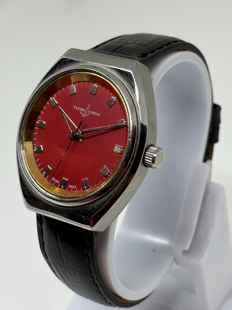 May include: A Ulysse Nardin wristwatch featuring a vibrant red dial with silver-tone hour markers and hands. The watch has a polished silver-tone metal case, a textured black leather strap, and the text Ulysse Nardin and Swiss Made on the dial.