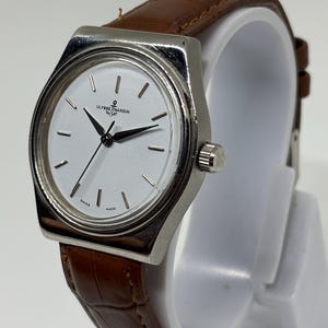 May include: A wristwatch with a silver-toned metal case and a white dial featuring black hands and hour markers. The dial displays the text Ulysse Nardin and Swiss Made. The watch is fitted with a brown leather strap with a textured crocodile pattern.