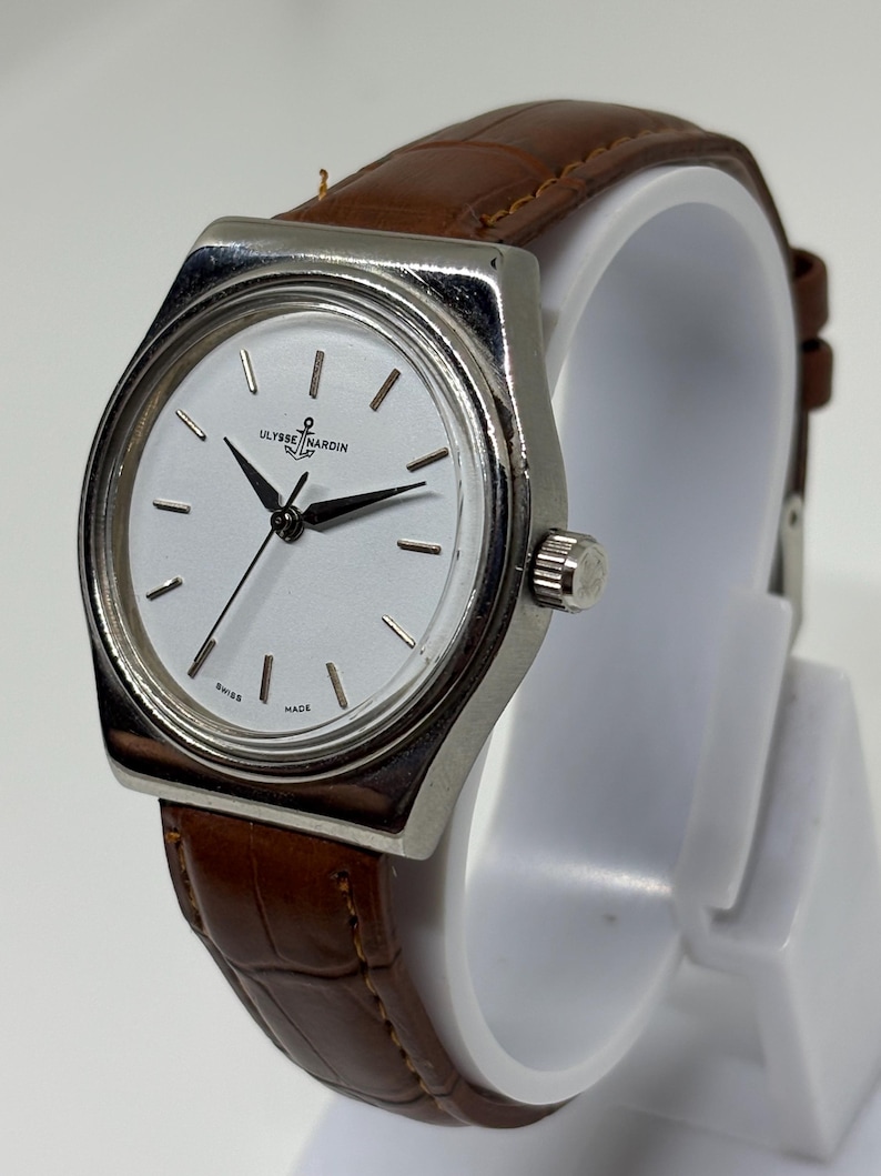 May include: A wristwatch with a silver-toned metal case and a white dial featuring black hands and hour markers. The dial displays the text Ulysse Nardin and Swiss Made. The watch is fitted with a brown leather strap with a textured crocodile pattern.