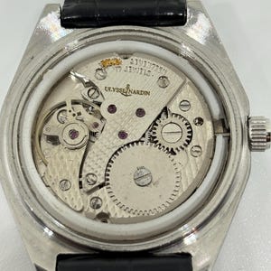 May include: A silver-toned Ulysse Nardin mechanical watch movement with an exposed back, featuring the text 'ULYSSE NARDIN' and 'SEVENTEEN 17 JEWELS' engraved on the metal plate, paired with a black leather strap with white stitching.