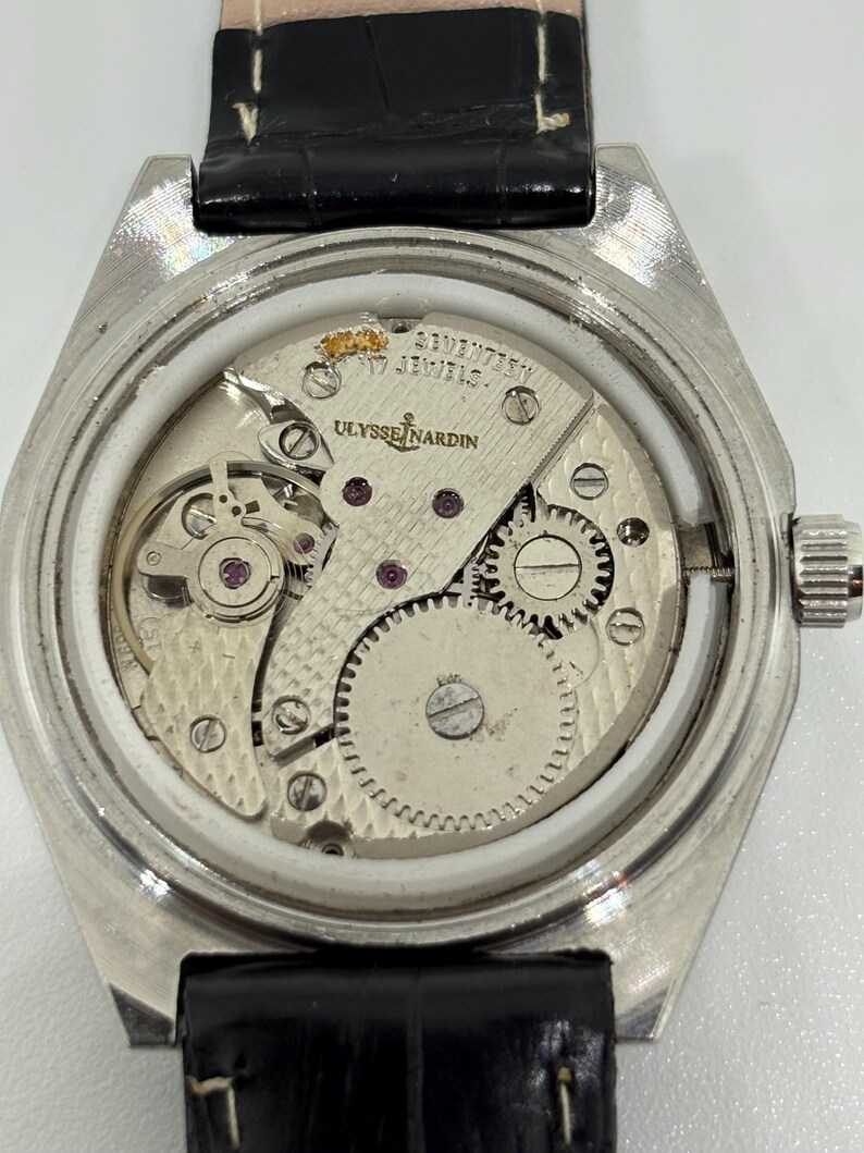 May include: A silver-toned Ulysse Nardin mechanical watch movement with an exposed back, featuring the text 'ULYSSE NARDIN' and 'SEVENTEEN 17 JEWELS' engraved on the metal plate, paired with a black leather strap with white stitching.